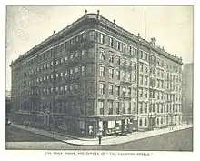 Black and white photo of an urban six-story Victorian commercial building of brick and cast iron on a street corner. The facade to the left is 23 bays wide, to the right is 18, plus a rounded corner one bay wide.