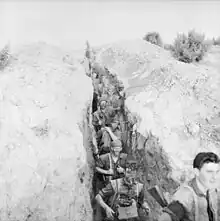 A deep trench line runs vertically from the top to the bottom of the photography, while on either side is a bare earth bund. Nine Caucasian soldiers carrying weapons, ammunition, and other equipment, many with their faces blacked and wearing camouflage on their heads, move in a straight line down the trench towards the bottom of the photo, below the camera. At the bottom right of the photo, a soldier stands level with the camera facing the right at the head of the patrol.