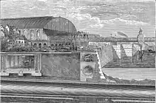 Illustrated section showing below ground structures of a riverside embankment including a covered railway line with steam train, riverside wall with sewer and pipes running behind. A large, glass roofed railway station sits in the left middle distance adjoining a railway bridge that crosses the river. Boats ply the water and tiny figures are engaged in construction work with a raised scaffold in the centre.