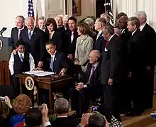 A man in a suit and tie sits at a wooden desk with two pieces of paper spread out in front of him. Behind him is a group of similarly dressed men and women; he is flanked by two young boys
